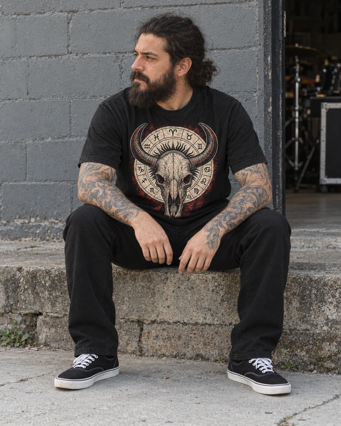 Un hombre con barba y brazos tatuados lleva la camiseta Cráneo, sentado junto a una pared gris